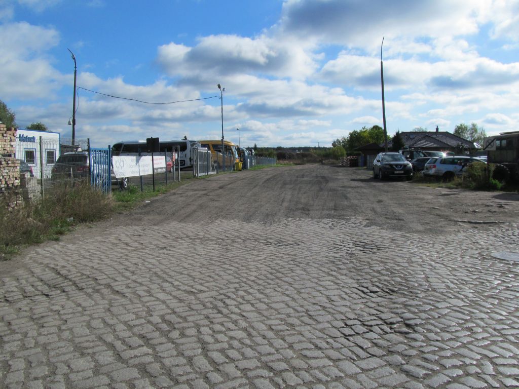 Ulica częściowo, na pierwszym planie wyłożona kamieniami, a w dalszej części staje się drogą żwirowa. Po dwóch stronach widać częściowo zabudowania i zaparkowane auta.