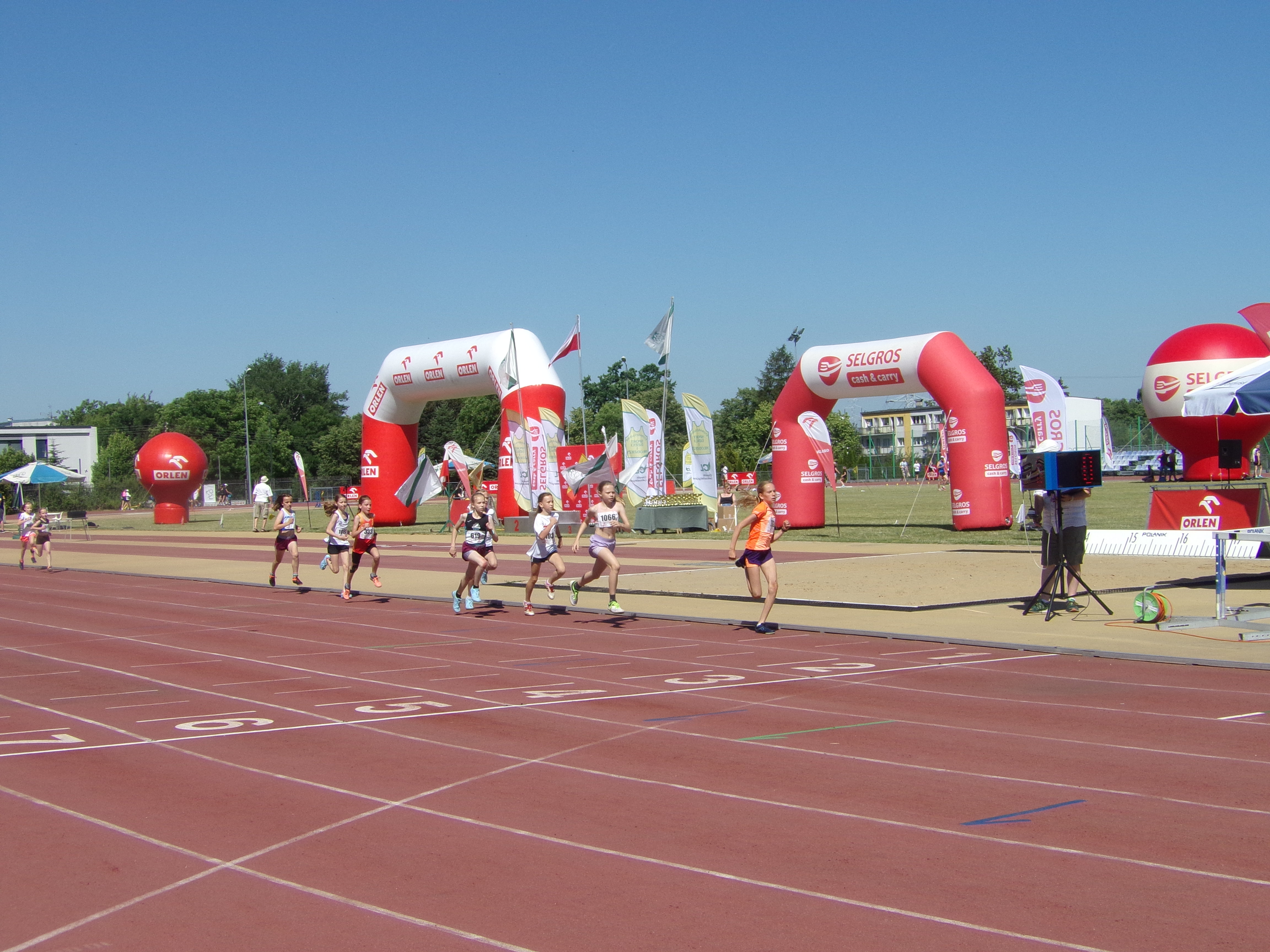 Po bieżni lekkoatletycznej biegnie dzisięć dziewczynek. W tle widać balony i flagi reklamowe.