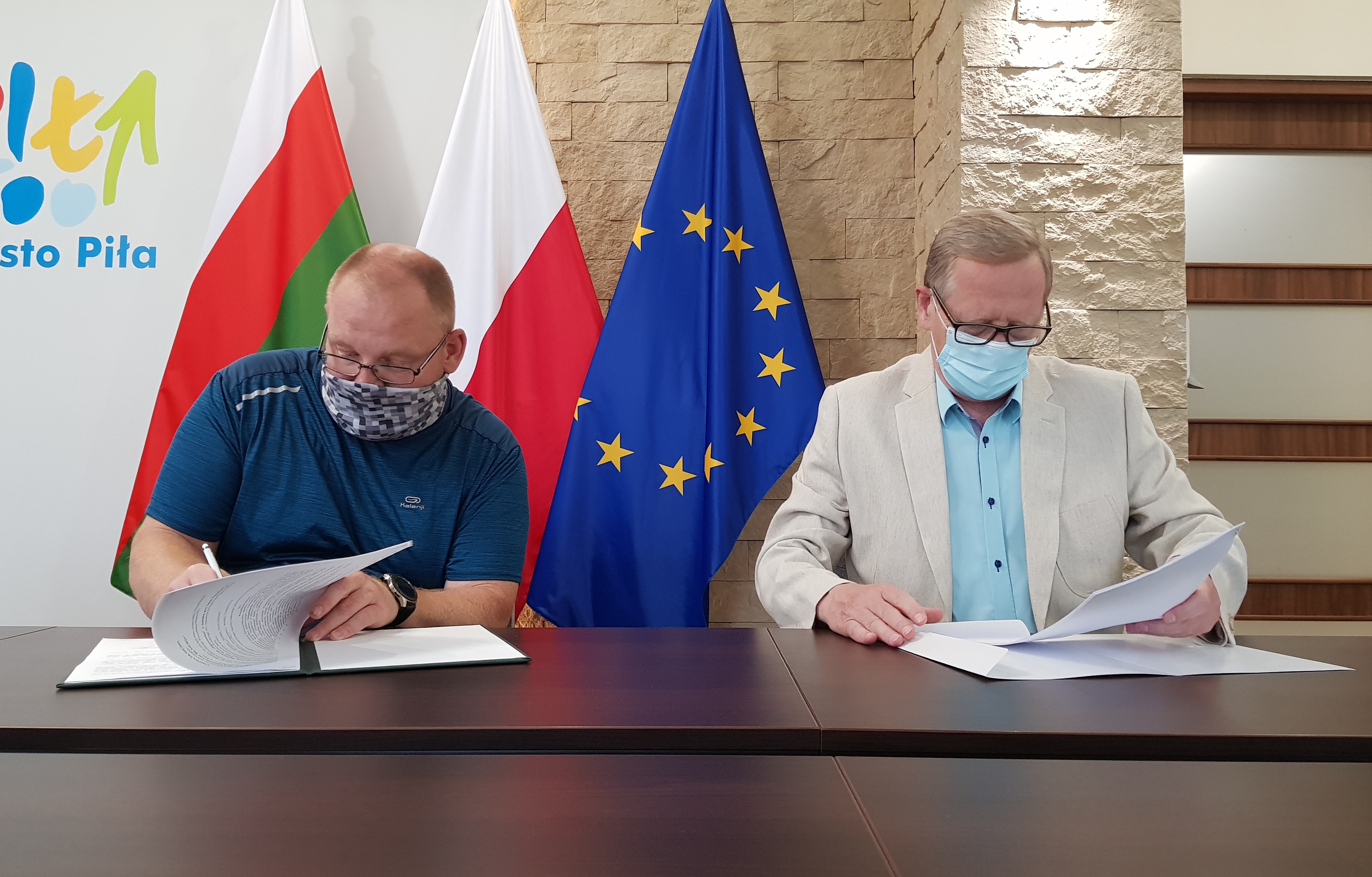 Dwóch mężczyzn siedzi przy stole, podpisują dokumenty. Znajdują się w pomieszczeniu, w tle stoją 3 flagi - miasta Piły, Polska i Unii Europejskiej. 