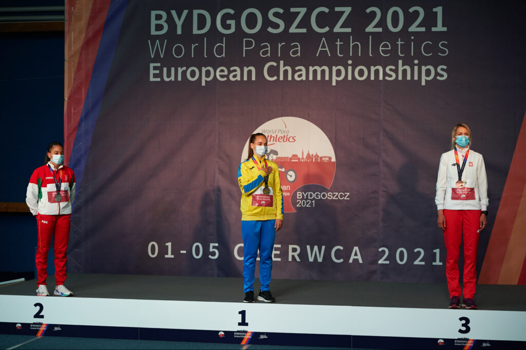 Trzy zawodniczki stoją na podium. Na szyjach mają zawieszone zdobyte medale. W tle widać baner z nazwą wydarzenia - Mistrzostwa Europy World Para Athletics, Bydgoszcz 2021. 