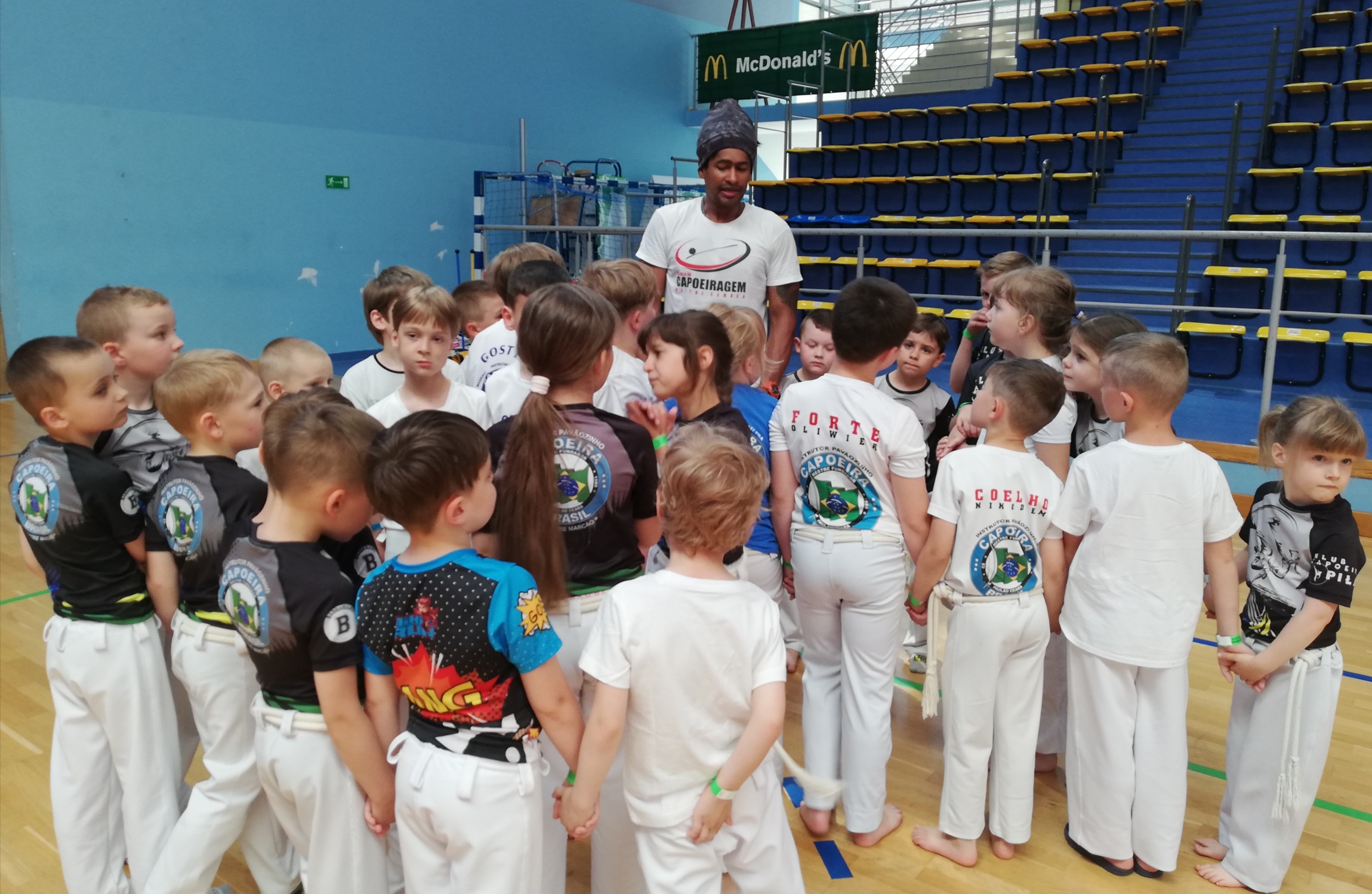 Instruktor capoeira stoi na sali sportowej w towarzystwie grupy dzieci i młodzieży. Młodzi sportowcy mają różne koszulki z logotypami klubów, które reprezentują. 