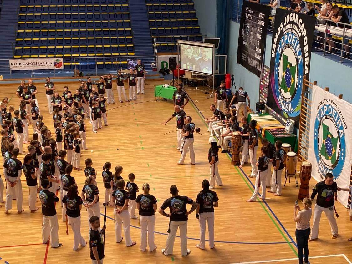 Hala sportowa. Po lewej stronie stoi duża grupa dzieci, po prawej instruktorzy capoeirą. Trenerzy pokazują ćwiczenia, dzieci i młodzież obserwują wykonywane ruchy. 