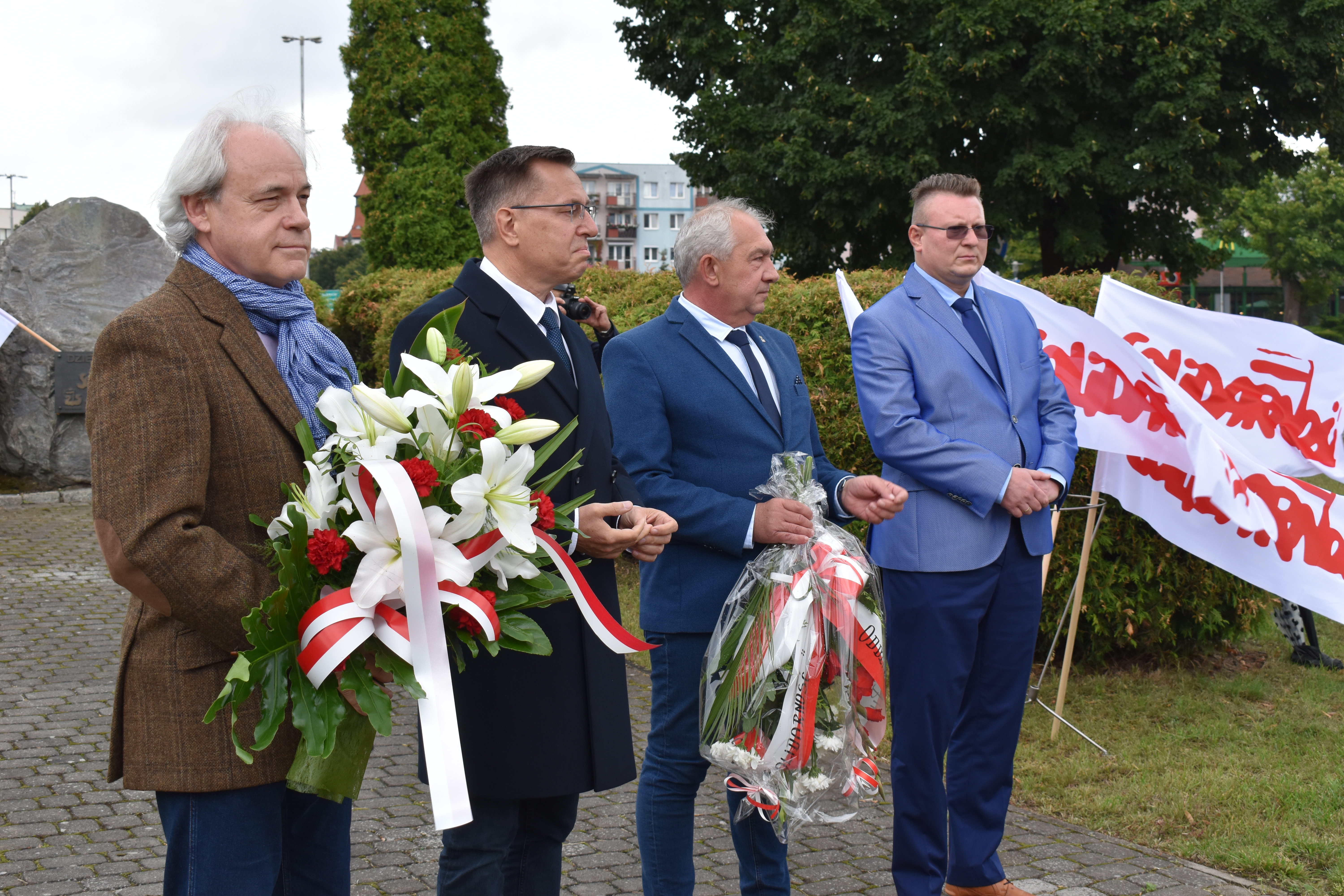 senator, prezydent, przewodniczący pilskeij Solidarności oraz wicestarosta stoją przed pomnikiem na rondzie Solidarności, po prawej stronie w koszu stoją flagi Solidarności
