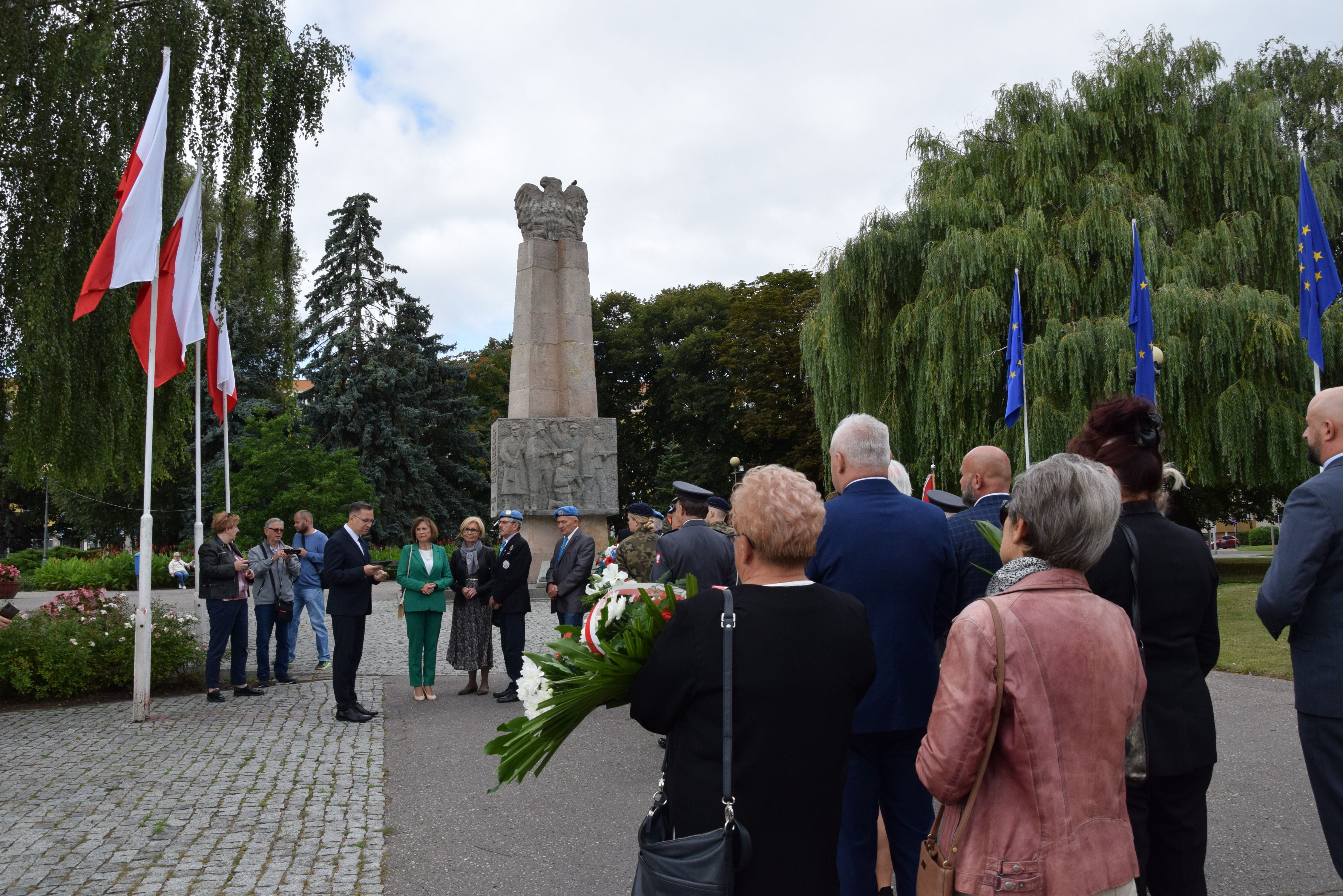 delegacje stoją przed pomnikiem, prezydent przemawia, w tle pomnik, po lewej flagi Polski