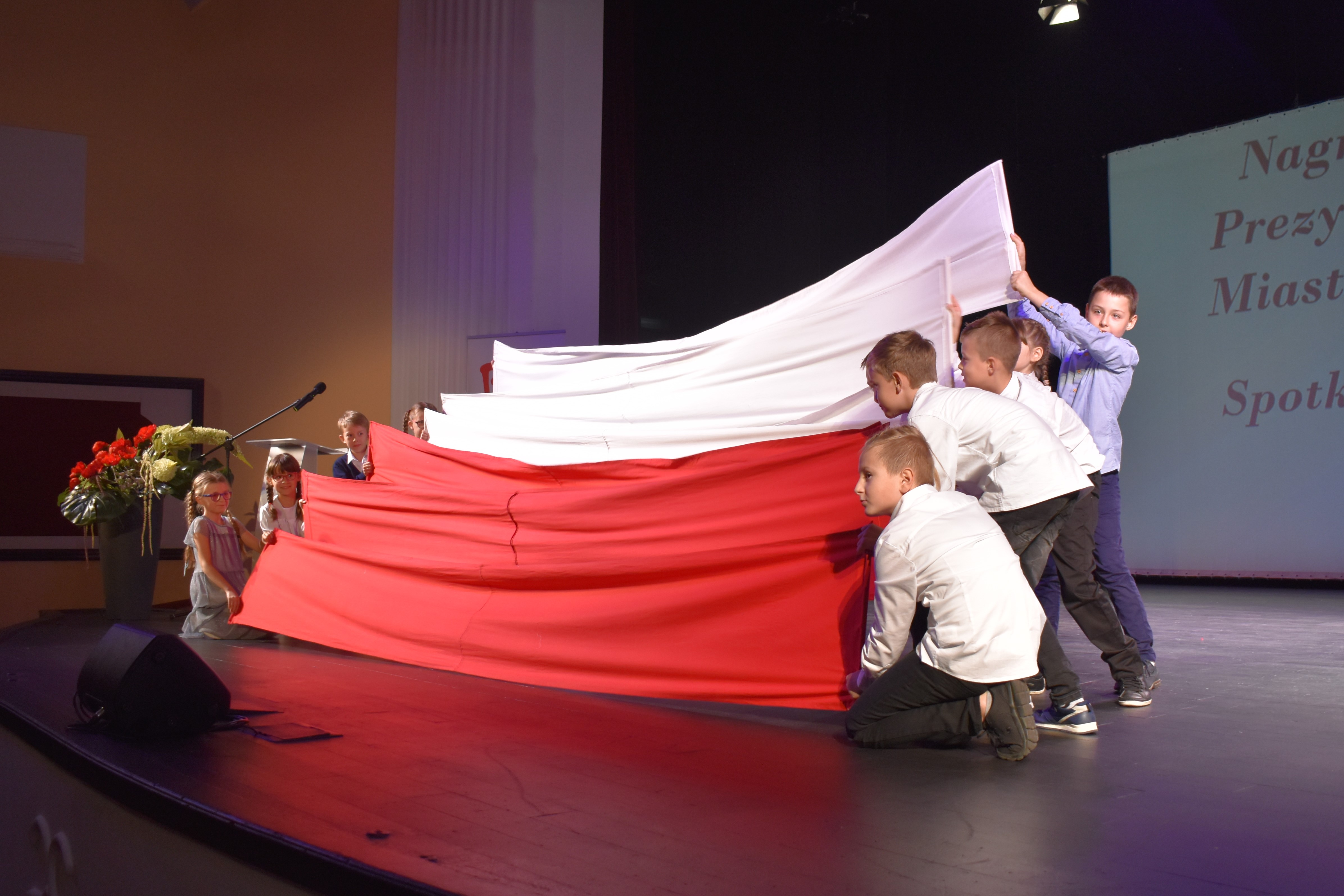 Dwanaścioro dziewczynek i chłopców stoi i kuca na scenie, trzymając z obu stron polskie flagi.
