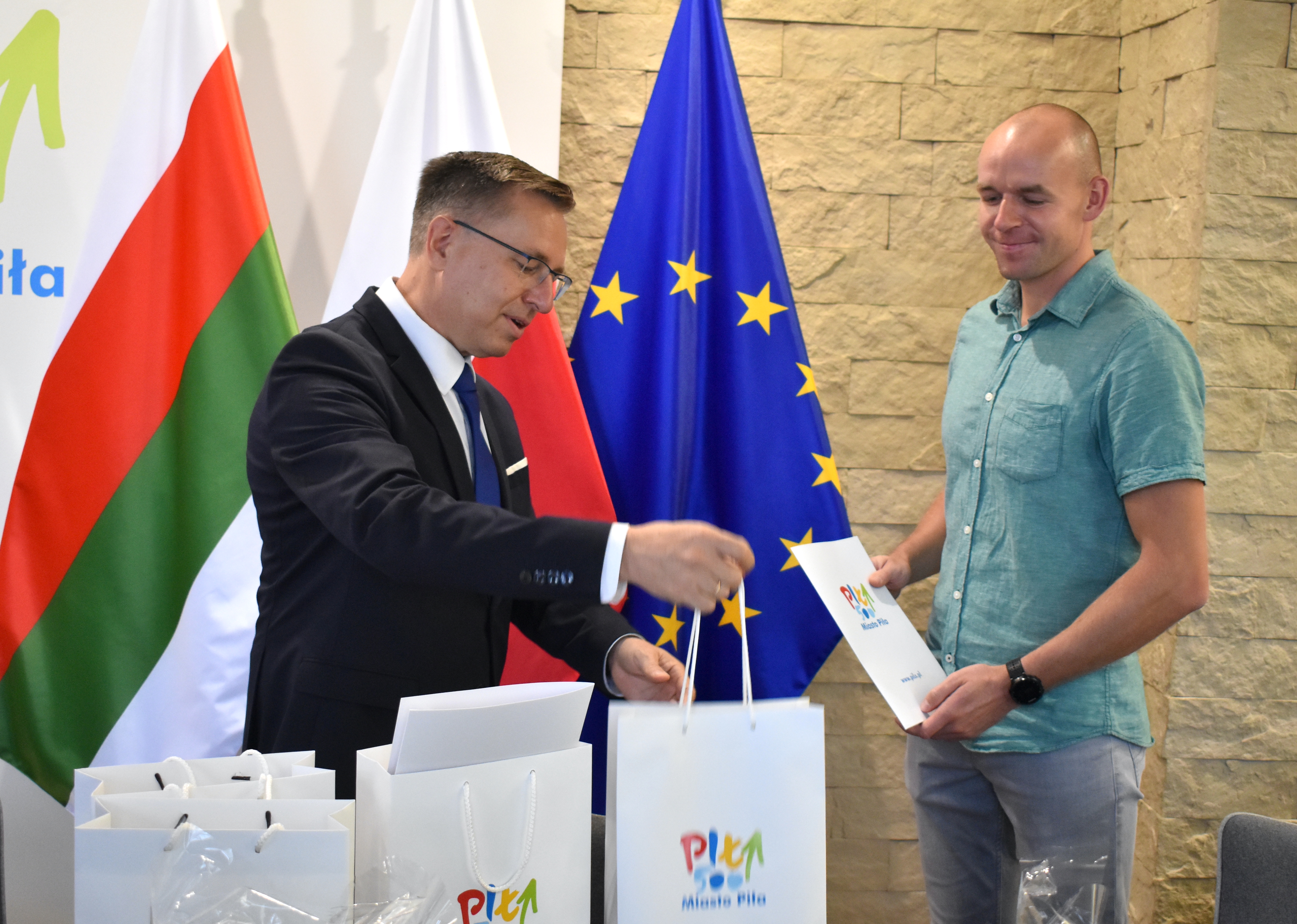 Dwóch mężczyzn, jeden trzyma w rękach teczkę, drugi wręcza paczkę. Na teczce i paczce widać logotyp miasta Piły. W tle stoją flagi miejska, Polska i Unii Europejskiej. 