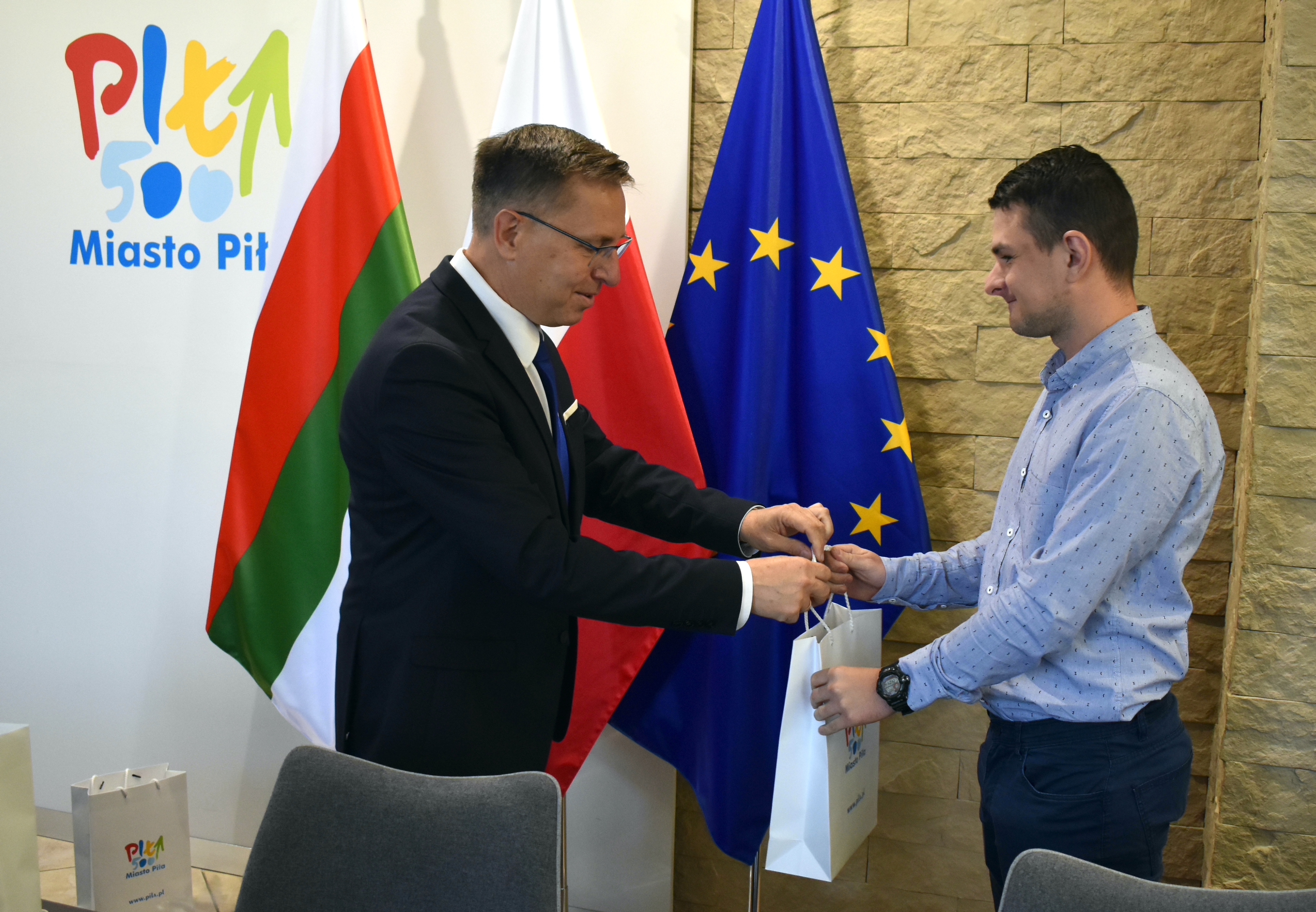 Dwóch mężczyzn - jeden wręcza drugiemu paczkę z upominkiem.  W tle widać logo miasta Piły, stoją flagi miejska, Polska i Unii Europejskiej. 